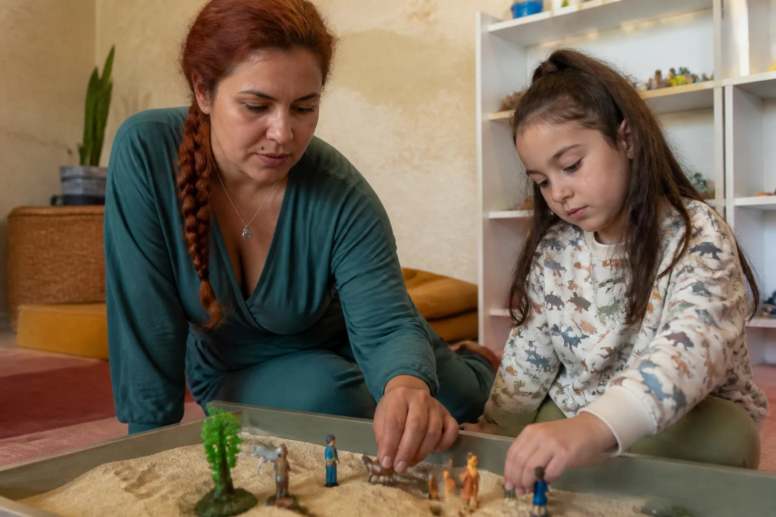 Oyun terapisi sırasında kum tepsisine küçük oyuncak figürler yerleştiren bir çocuk ve psikolog